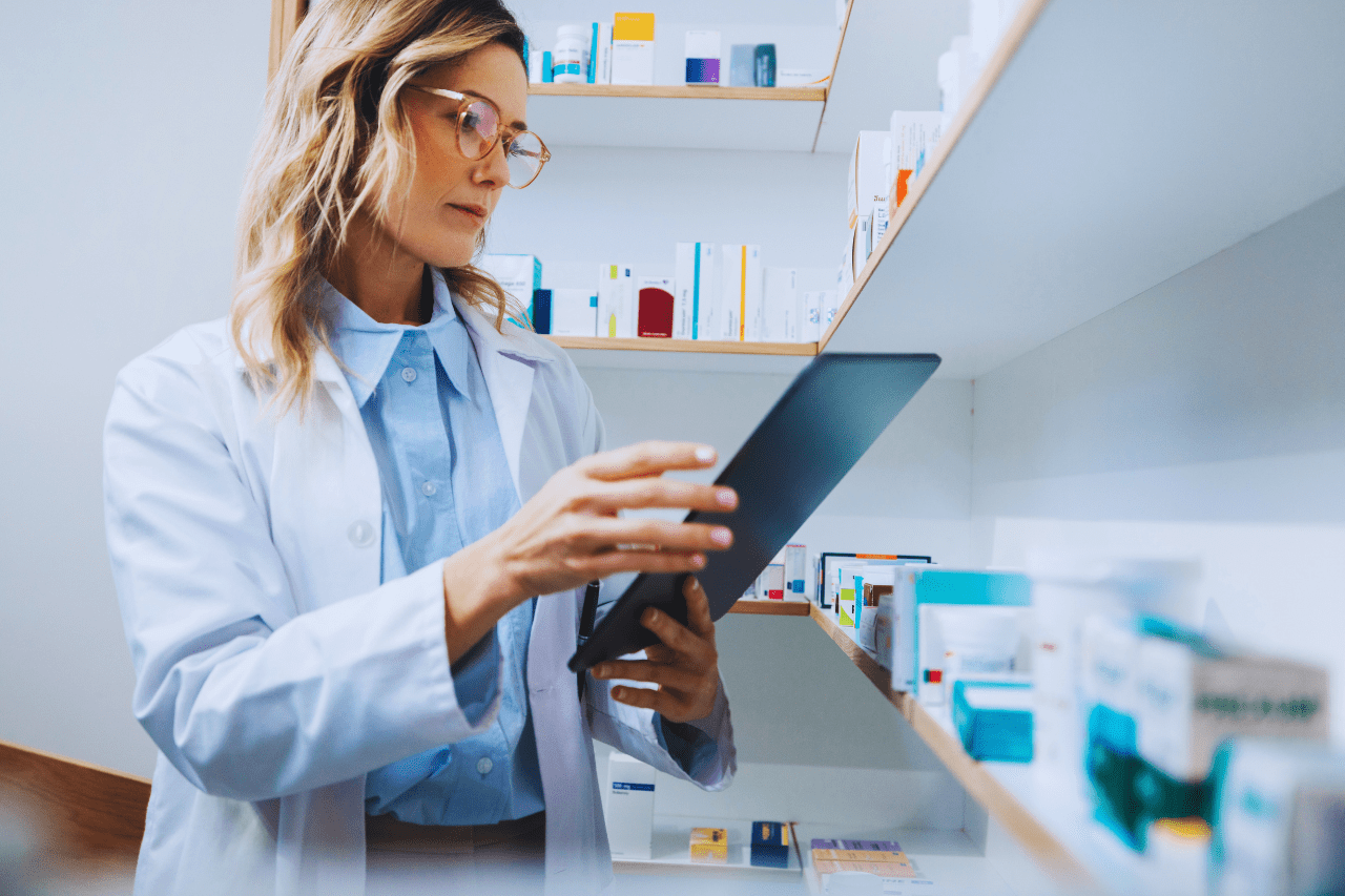 Pharmacist in white coat using a tablet while standing near shelves of medications