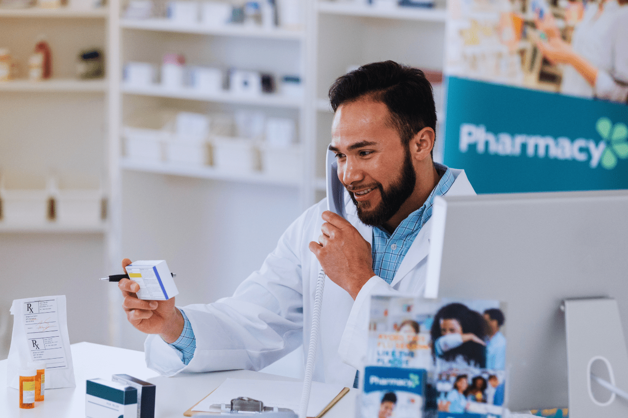 Pharmacist in white coat examining a medication box at a pharmacy counter with shelves in background